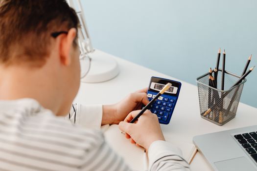Close-up of a person calculating numbers at a desk with a pencil and laptop.