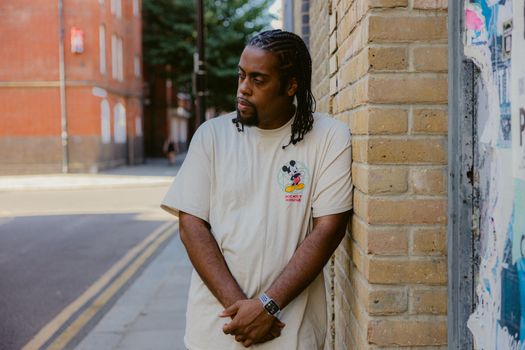A man with dreadlocks in a casual outfit stands against an urban brick wall.
