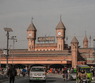 A bustling scene outside a historic railway station featuring prominent clock towers and Arabic signage.