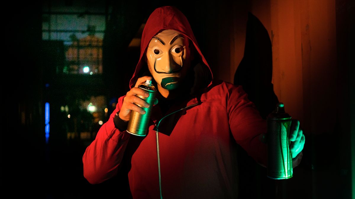A masked person in a red hoodie holds spray paints, creating street art at night.