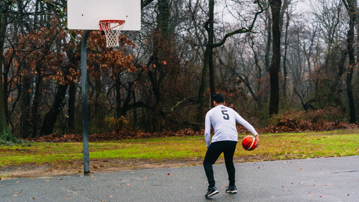 Adult man playing basketball on an outdoor court surrounded by autumn trees.