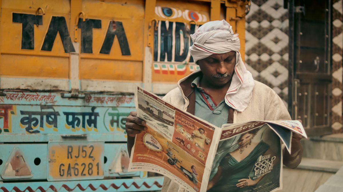 Man with mustache reads a newspaper next to a Tata dump truck on an urban street.
