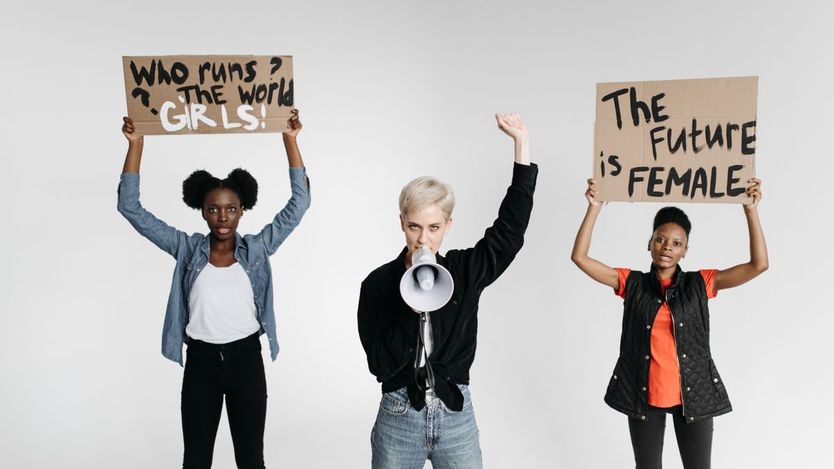Three women holding protest signs and a megaphone advocating for women's rights in a studio setup.