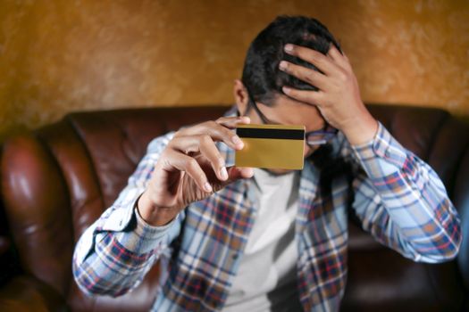 Man in checkered shirt on couch holding credit card, showing stress and worry.
