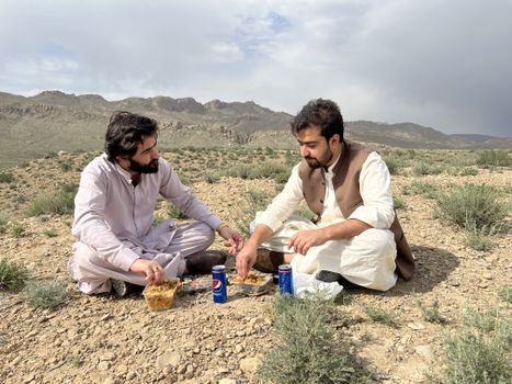 Two men sharing a meal outdoors in Quetta, Balochistan with scenic mountain views.