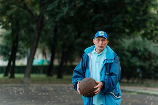 Senior man in blue jacket and cap holding basketball outdoors.