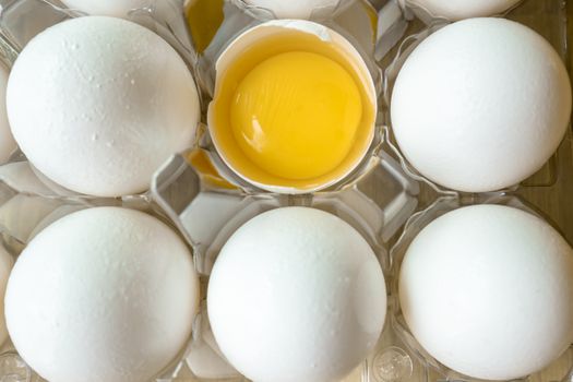 A high-angle view of fresh eggs in a carton with one cracked open showing the yolk.