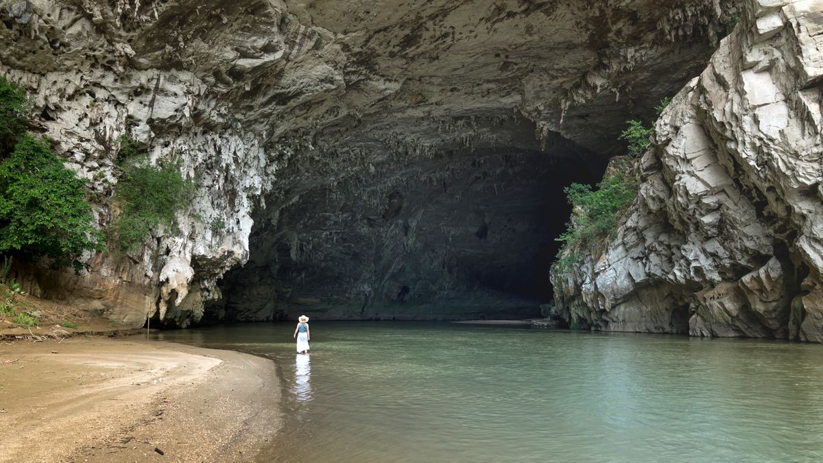A traveler stands at the entrance of a large, picturesque cave with a river flowing out.