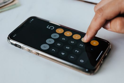 Crop faceless financier touching mobile phone screen during work while calculating on calculator application representing digits and financial symbols