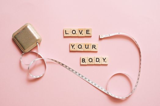 Scrabble letters spelling 'Love Your Body' with a tape measure on a pink backdrop.