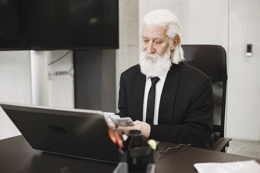White-haired man in suit counting money at a modern office desk with laptop.