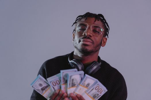 Portrait of a man holding bundles of cash wearing glasses and headphones. Indoor setting.